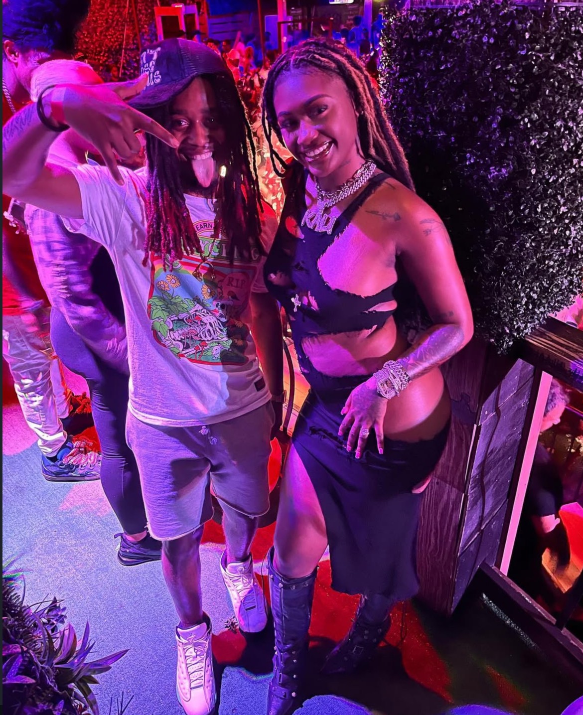 Two people smiling at a party, one with dreadlocks and the other with long hair, in colorful lighting.