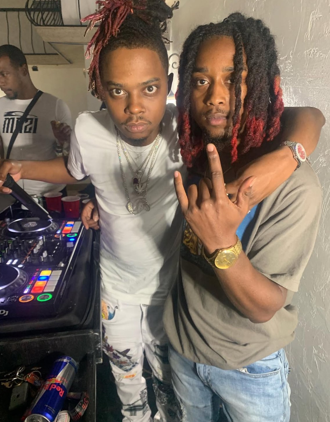 Two men pose together, one with dreadlocks and the other with red-tinted hair, in a room with a DJ setup nearby.