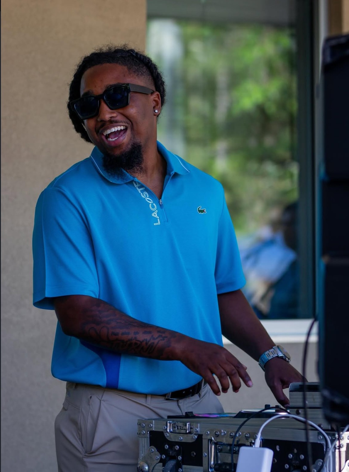 Man smiling, wearing sunglasses and a blue polo shirt, standing outdoors with greenery in the background.