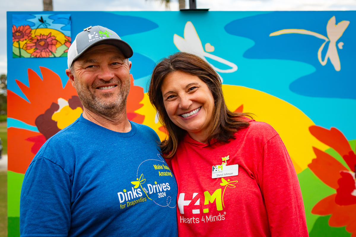 Two smiling people standing outdoors in front of colorful artwork, one wearing a blue shirt and the other a red shirt.