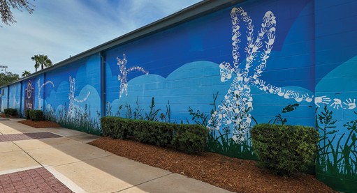 Colorful mural of a rabbit and plants on a blue wall, with a sidewalk and bushes in front.