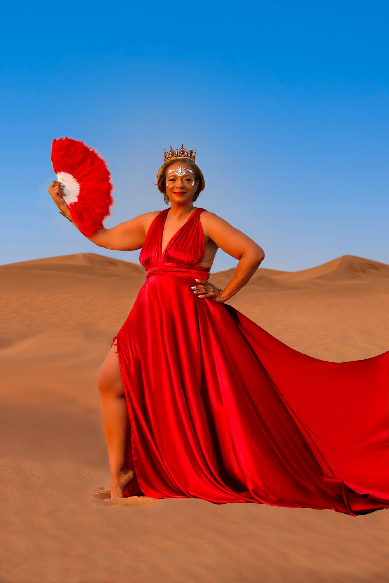 Woman in red dress holding fan, standing in desert with sand dunes and blue sky.