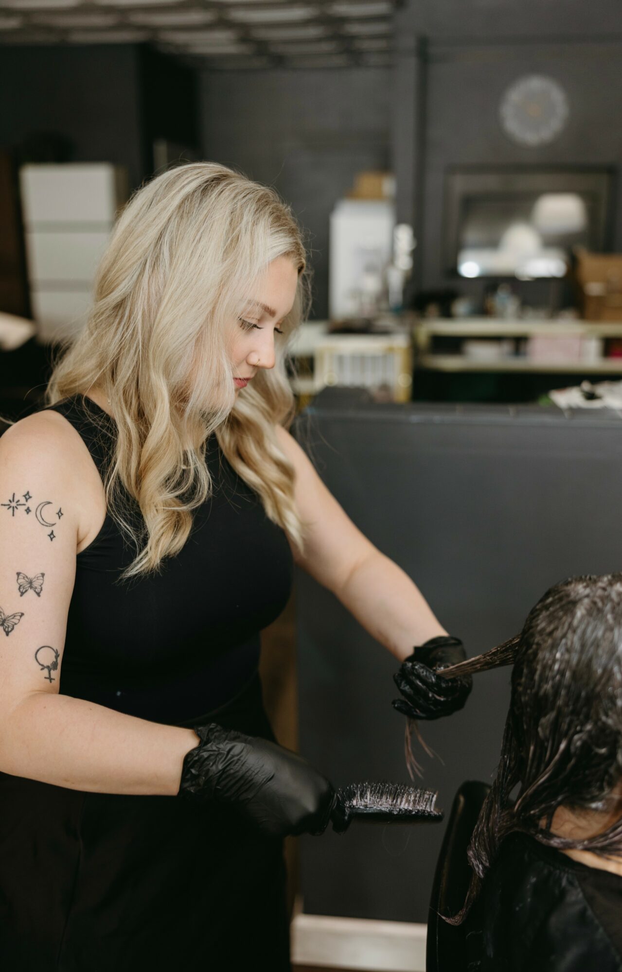 Woman with blonde hair applying hair dye to a person's hair in a salon, wearing black gloves.