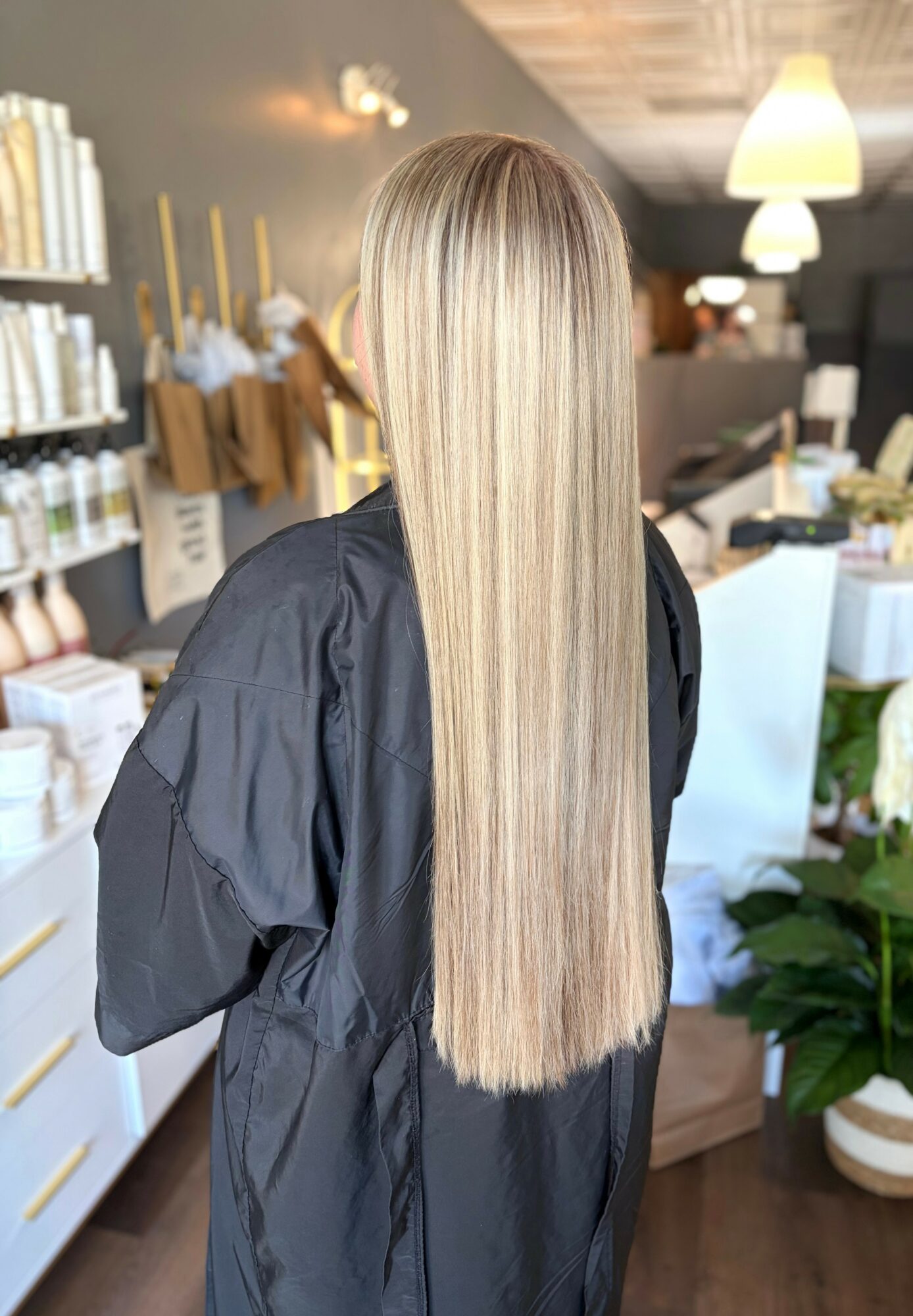 Person with long, straight, blonde hair in a salon, facing away, wearing a black cape, shelves with products behind.