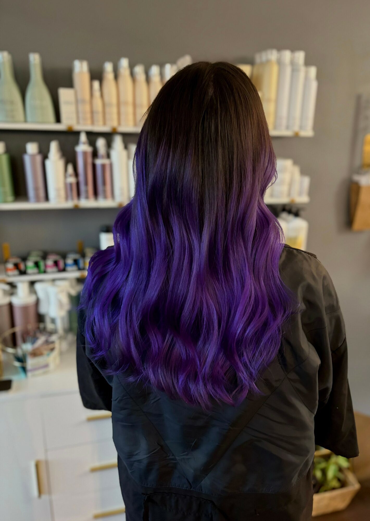 Person with long, wavy hair dyed in shades of purple and blue, standing in a store with shelves of bottles.