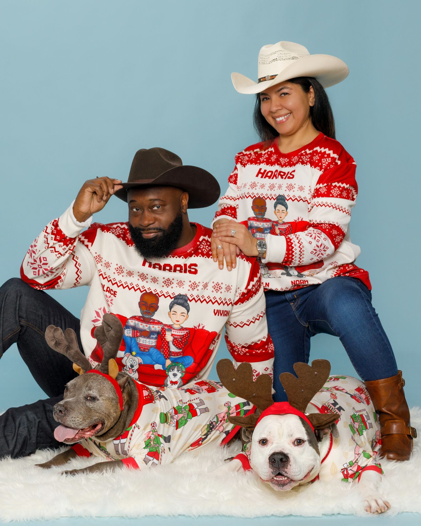 Two people wearing Christmas sweaters and cowboy hats, sitting with two dogs in reindeer costumes, against a blue background.