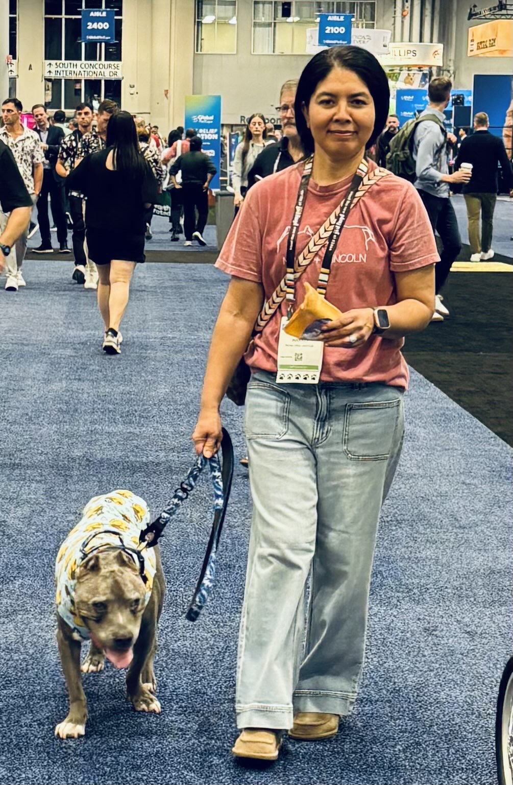 Woman walking with a dog on a leash in a busy indoor event space, holding a yellow card, smiling.