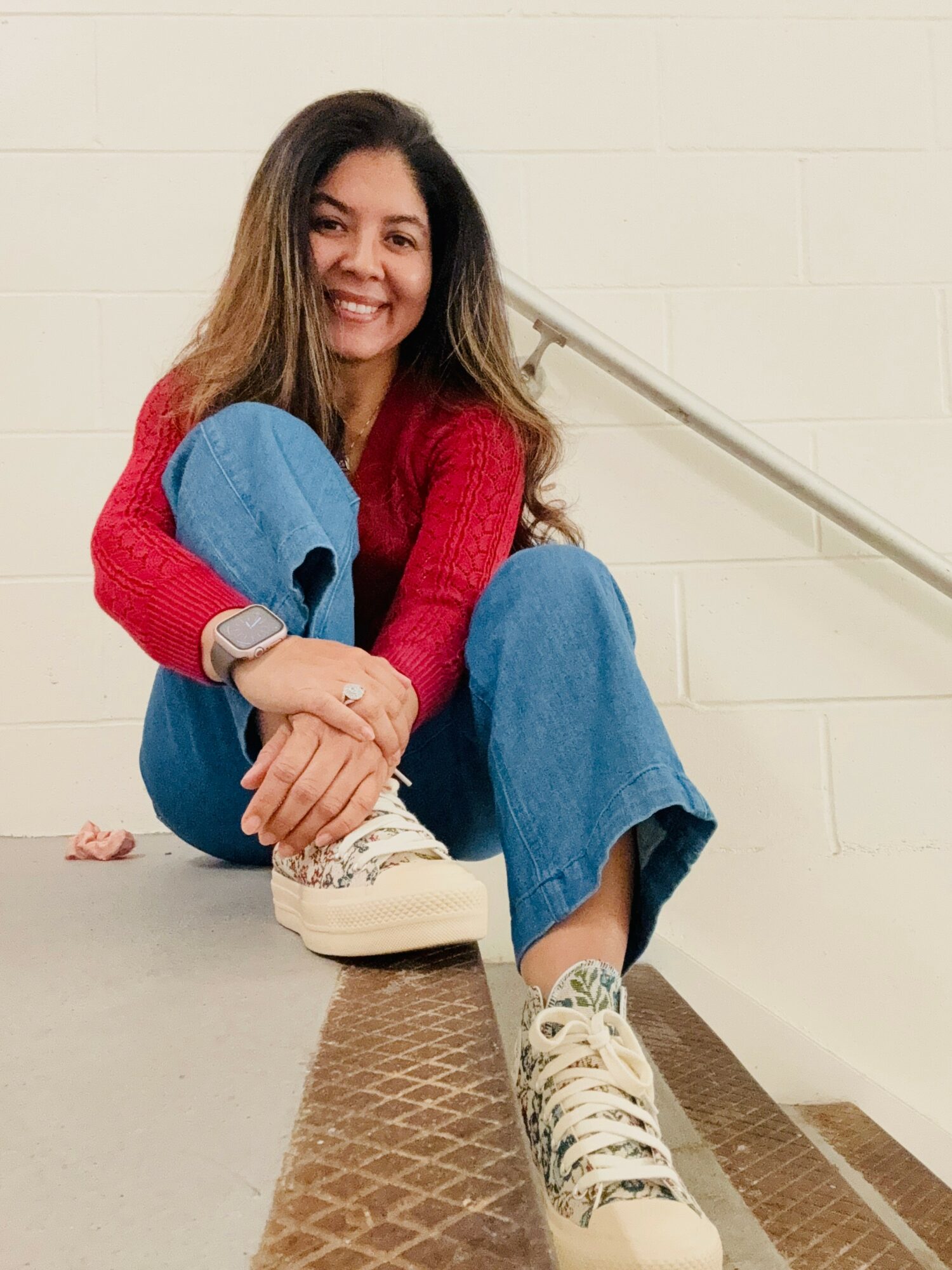 Woman sitting on stairs, smiling, wearing a red sweater, blue jeans, and sneakers, with a white brick wall background.