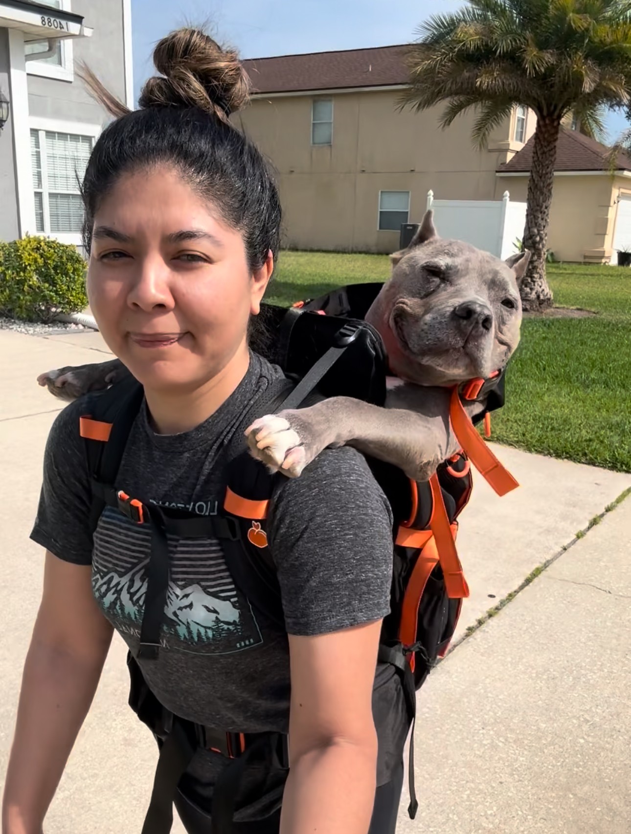 Young woman with dark hair in a bun carrying a dog in a backpack outdoors, with trees and buildings in background.