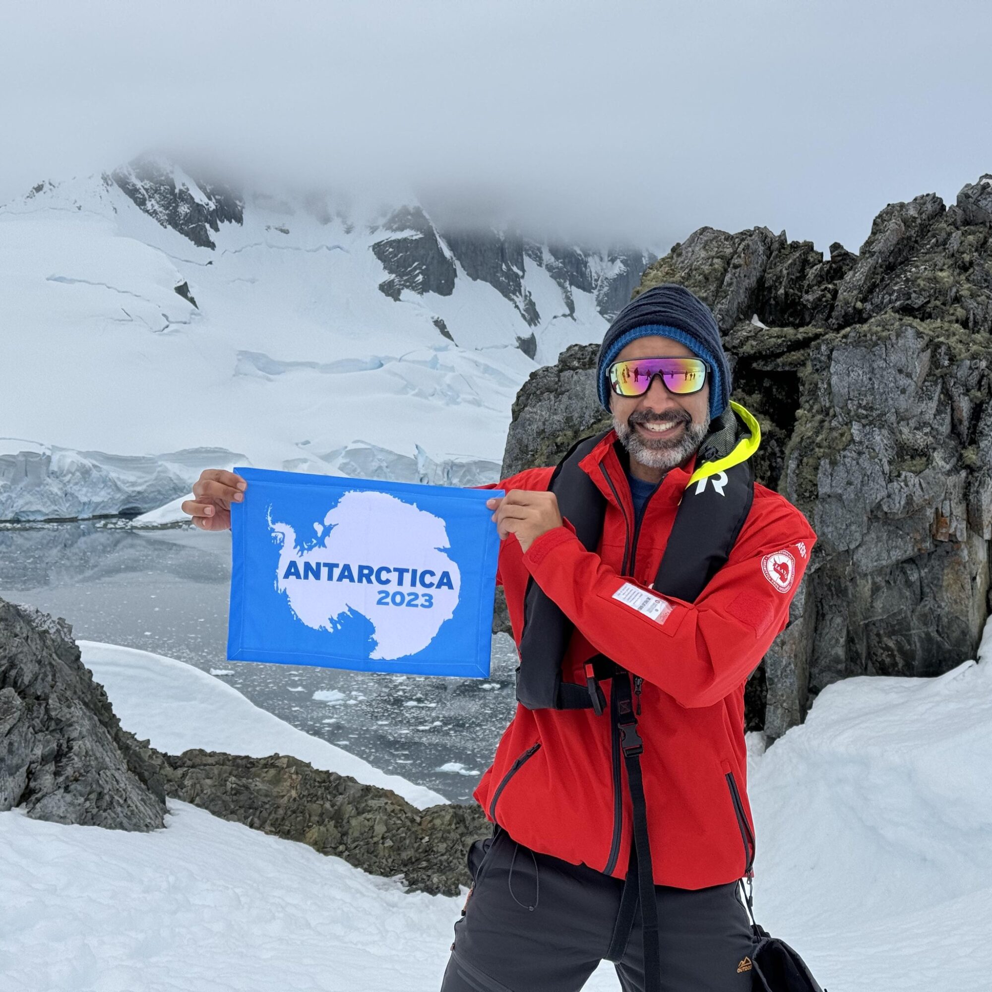 Man in red jacket holding a blue flag with Antarctica 2023, snowy mountain background.