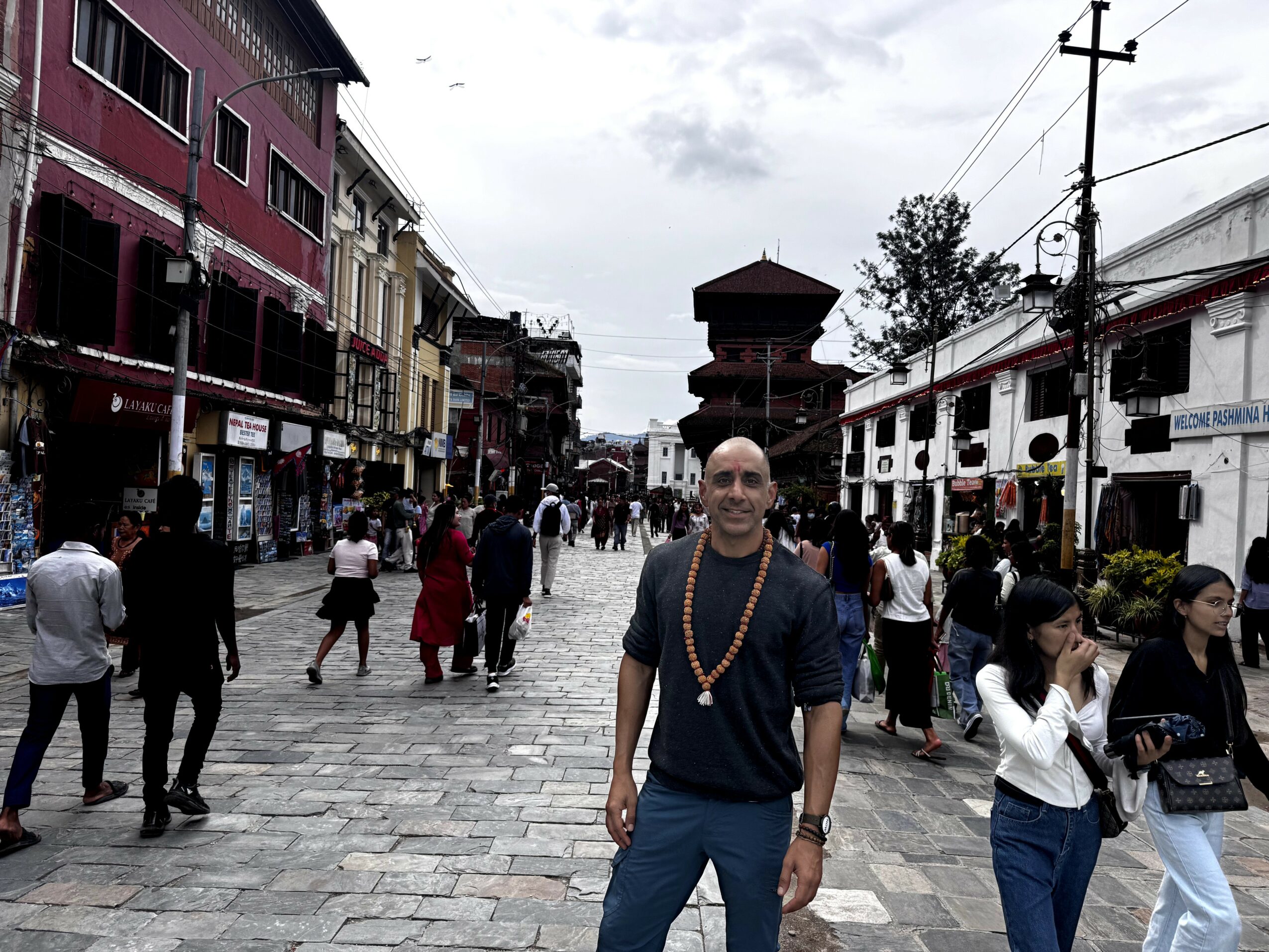 Bald man wearing a necklace standing on a busy cobblestone street with shops and people walking around.