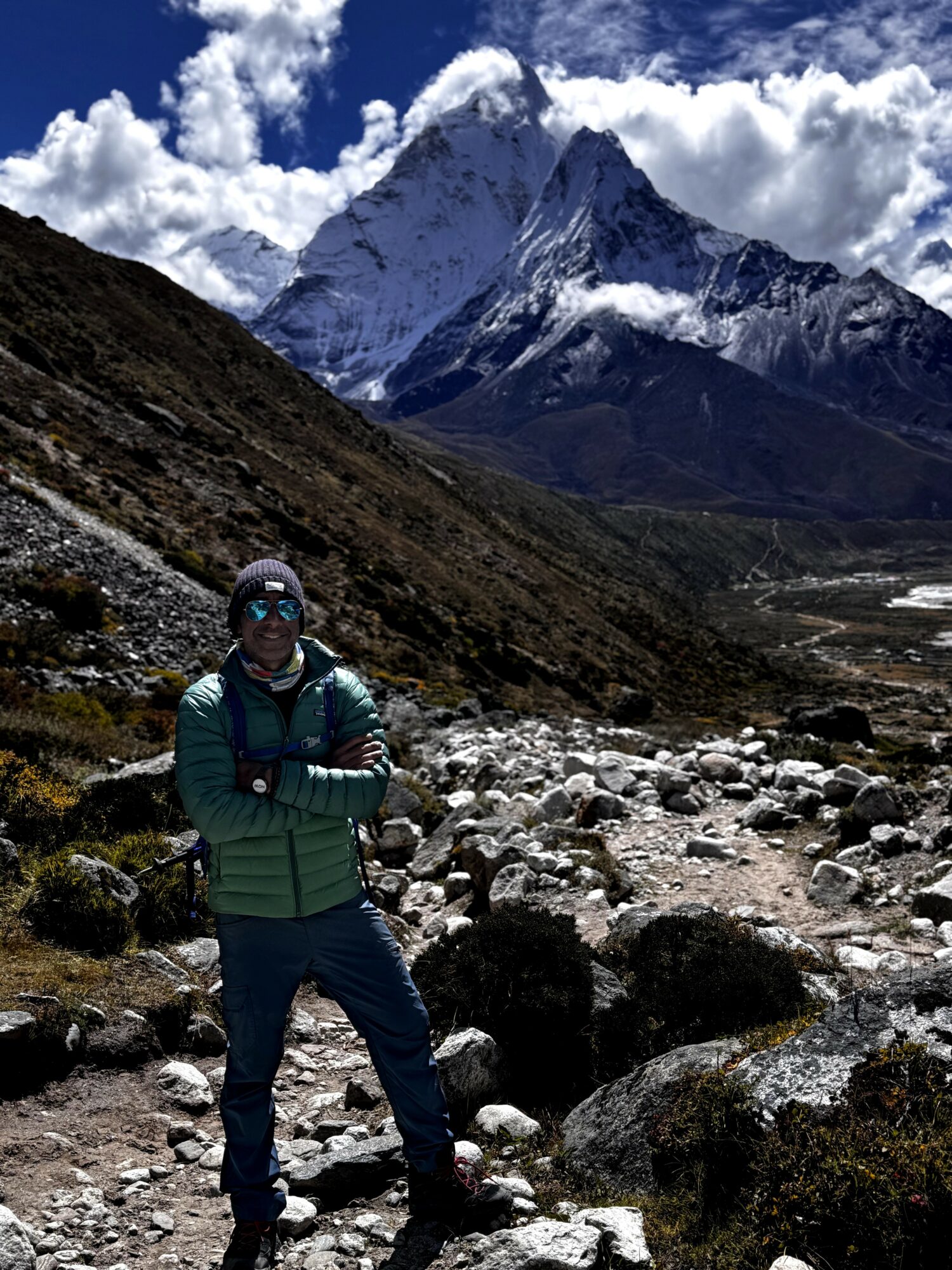 Person standing on rocky trail with snow-capped mountain and clouds in background.
