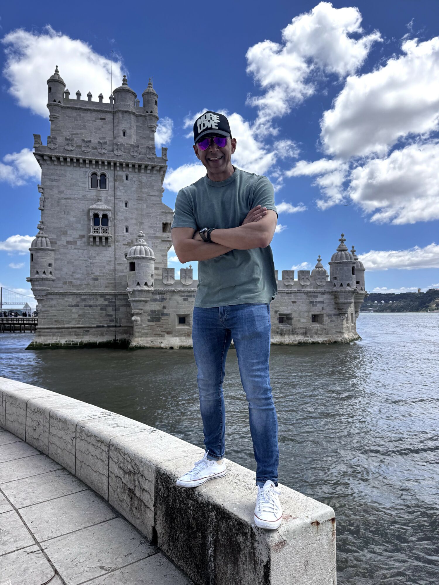 Person standing on a stone ledge in front of a historic castle by water, with a blue sky and clouds above.