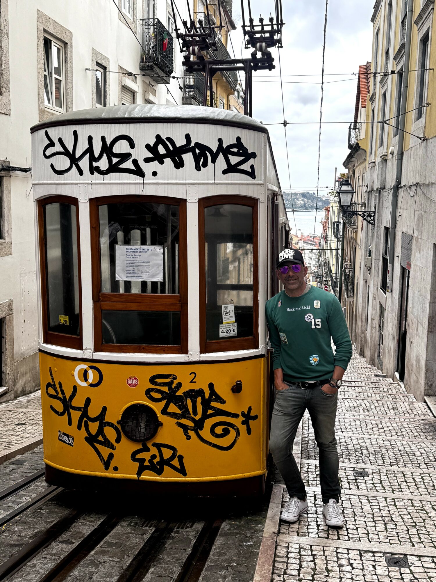 Person standing next to a yellow tram with graffiti, on a narrow street with buildings and overhead wires.