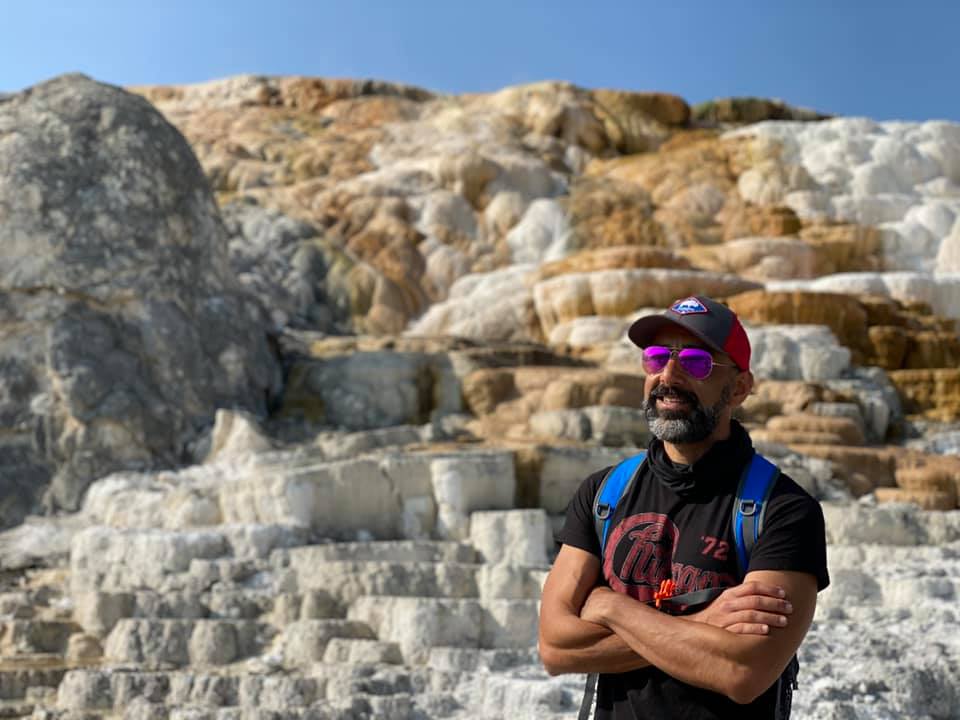 Man with sunglasses, beard, red cap, black shirt, and backpack standing on stone steps outdoors.