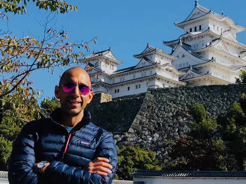 Man wearing sunglasses and a dark jacket standing outdoors with a traditional Japanese castle in the background.