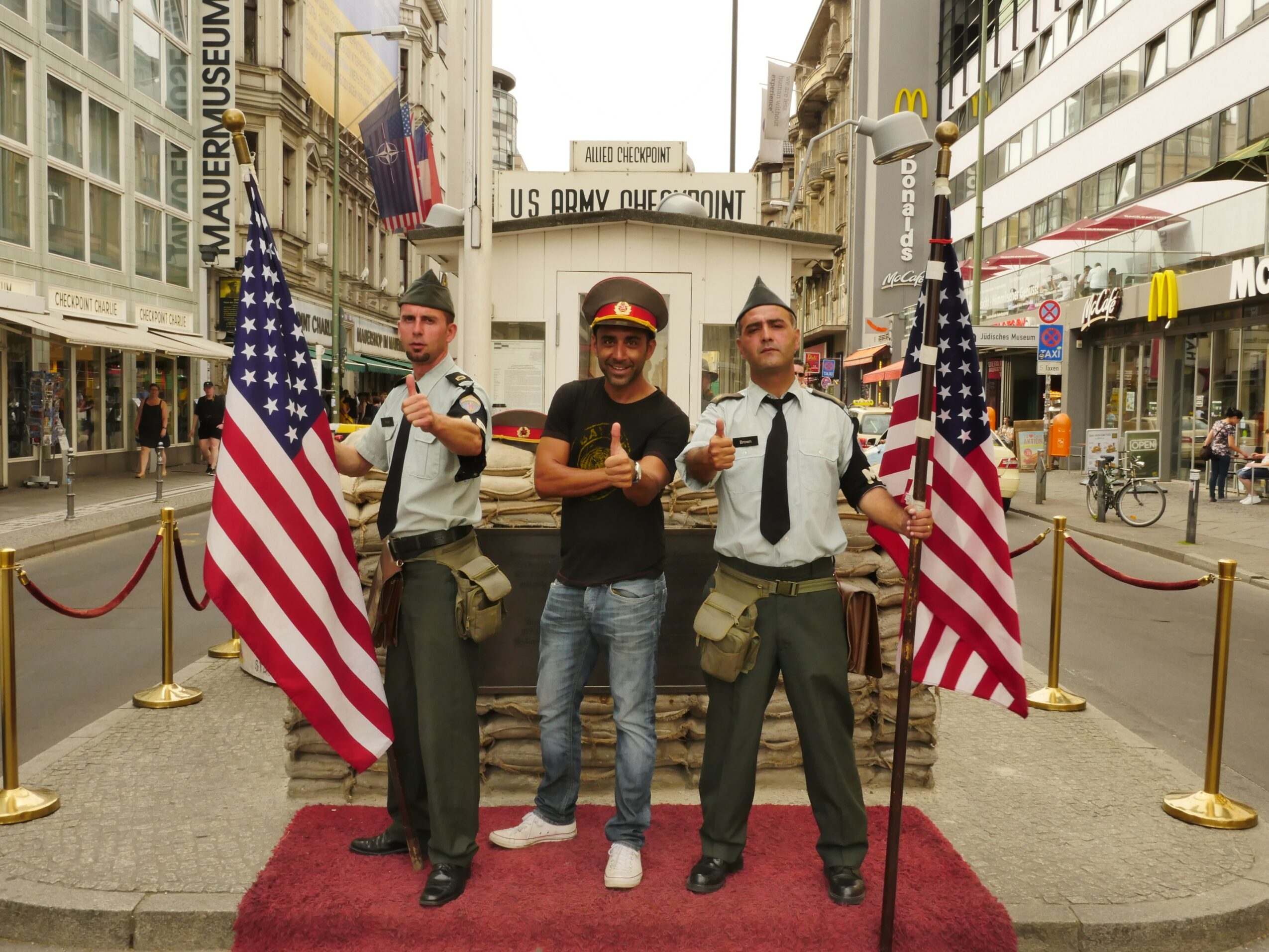 Three men stand on a red carpet in front of a historical building, flanked by American flags, in an urban setting.