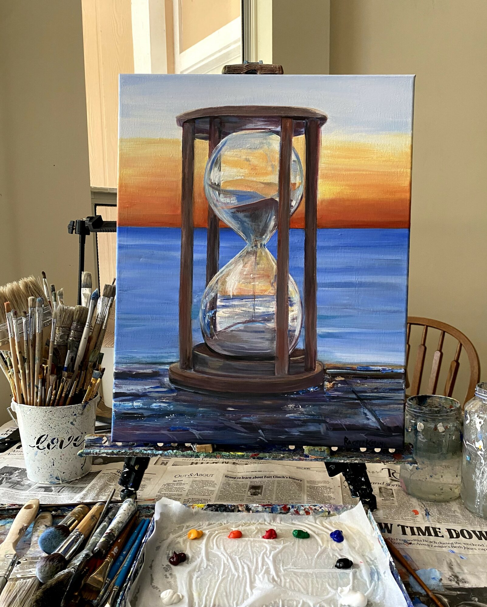 Painting of an hourglass on a wooden surface with a sunset and water background, in a room with art supplies.