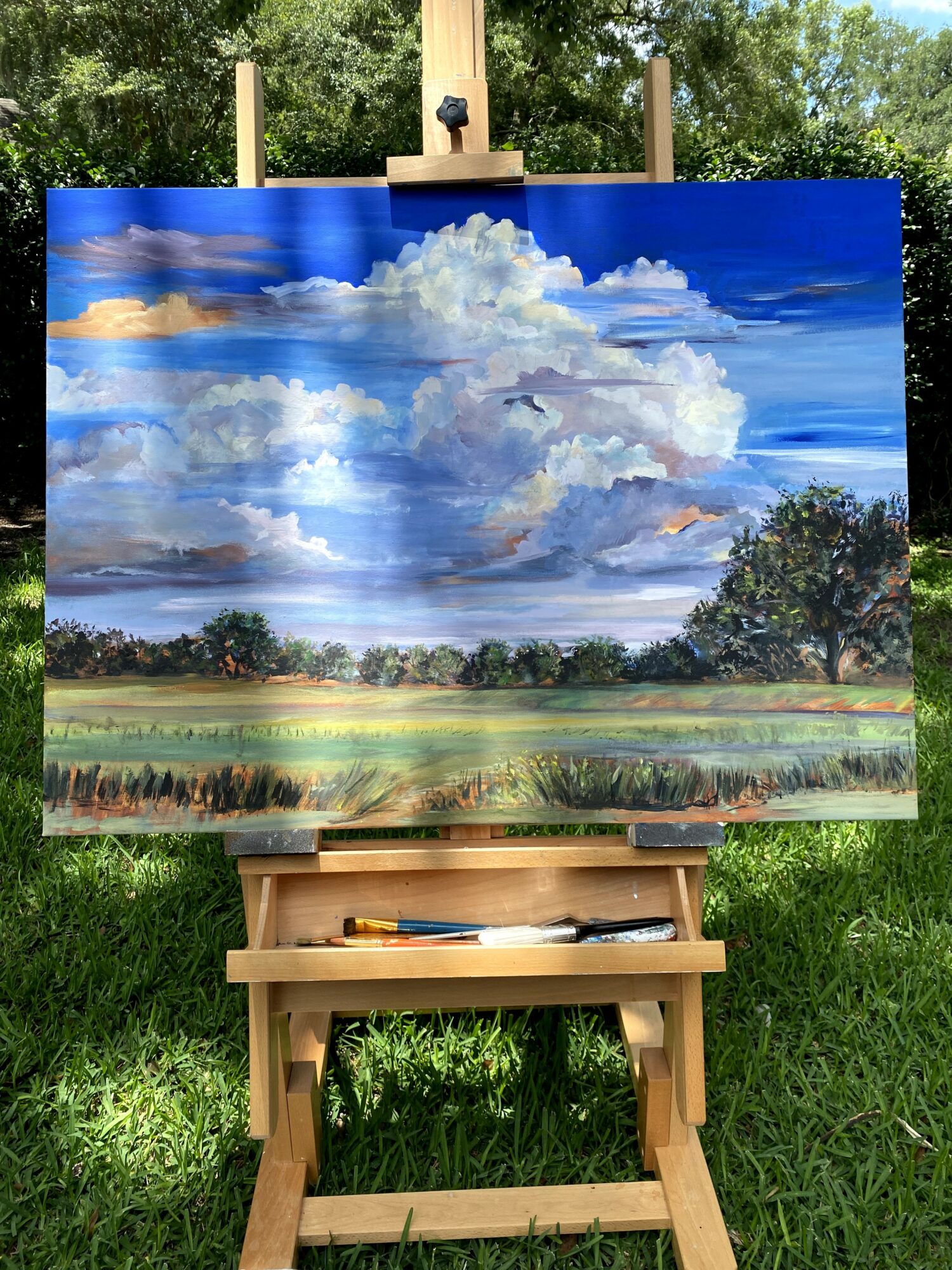 Landscape painting of a sky with clouds, trees, and grass, displayed outdoors on an easel.