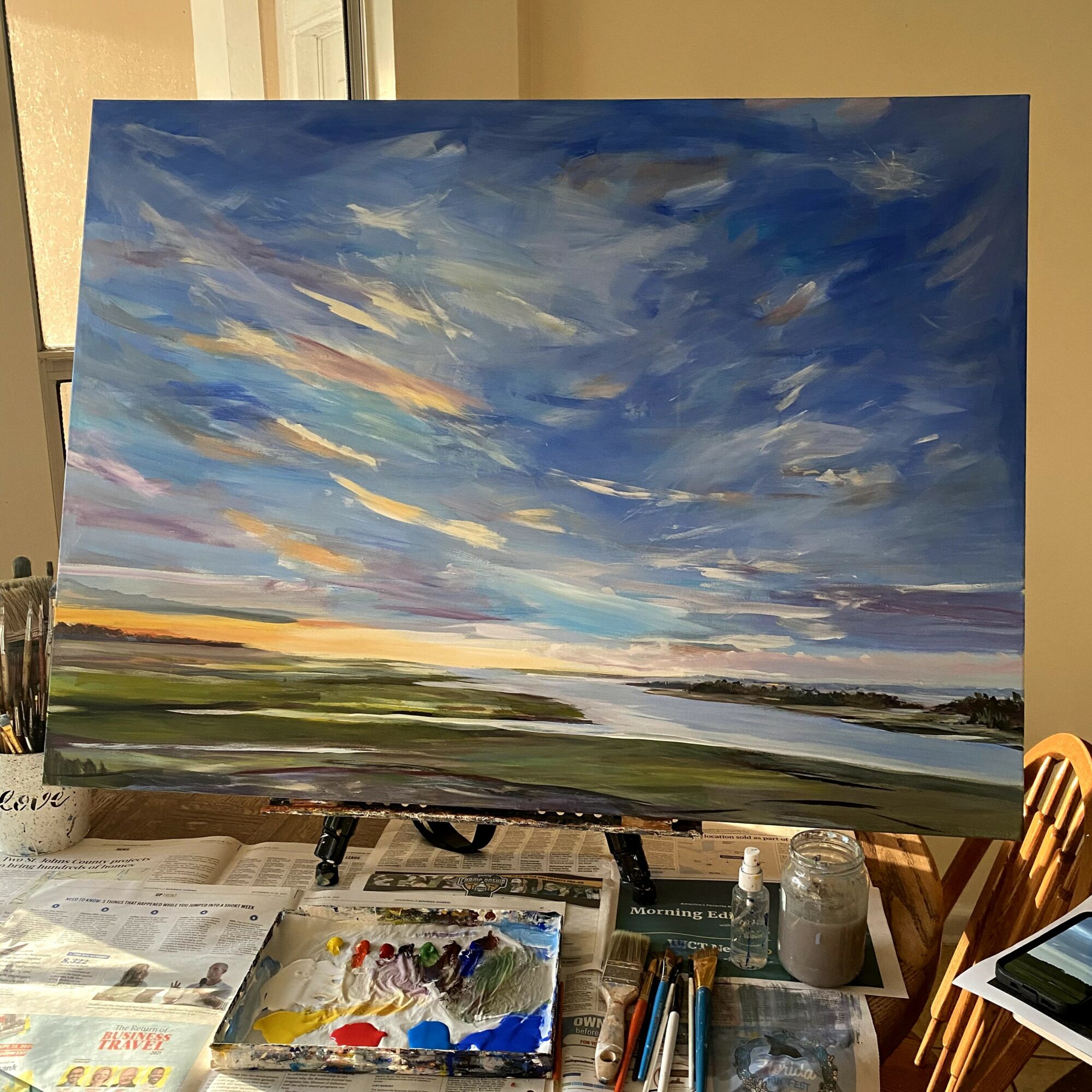 Landscape painting of a sky with clouds, a horizon, and a body of water, on an easel in a room.
