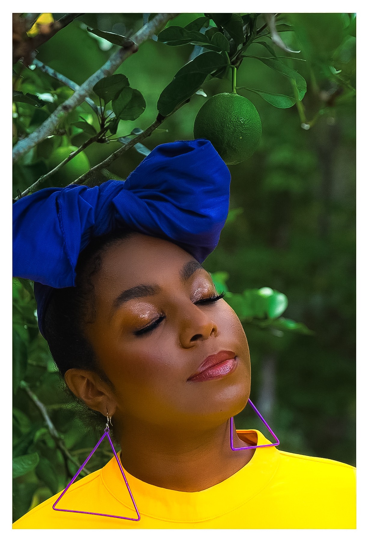 Woman with closed eyes, wearing a blue headwrap and yellow top, outdoors with green fruit and leaves