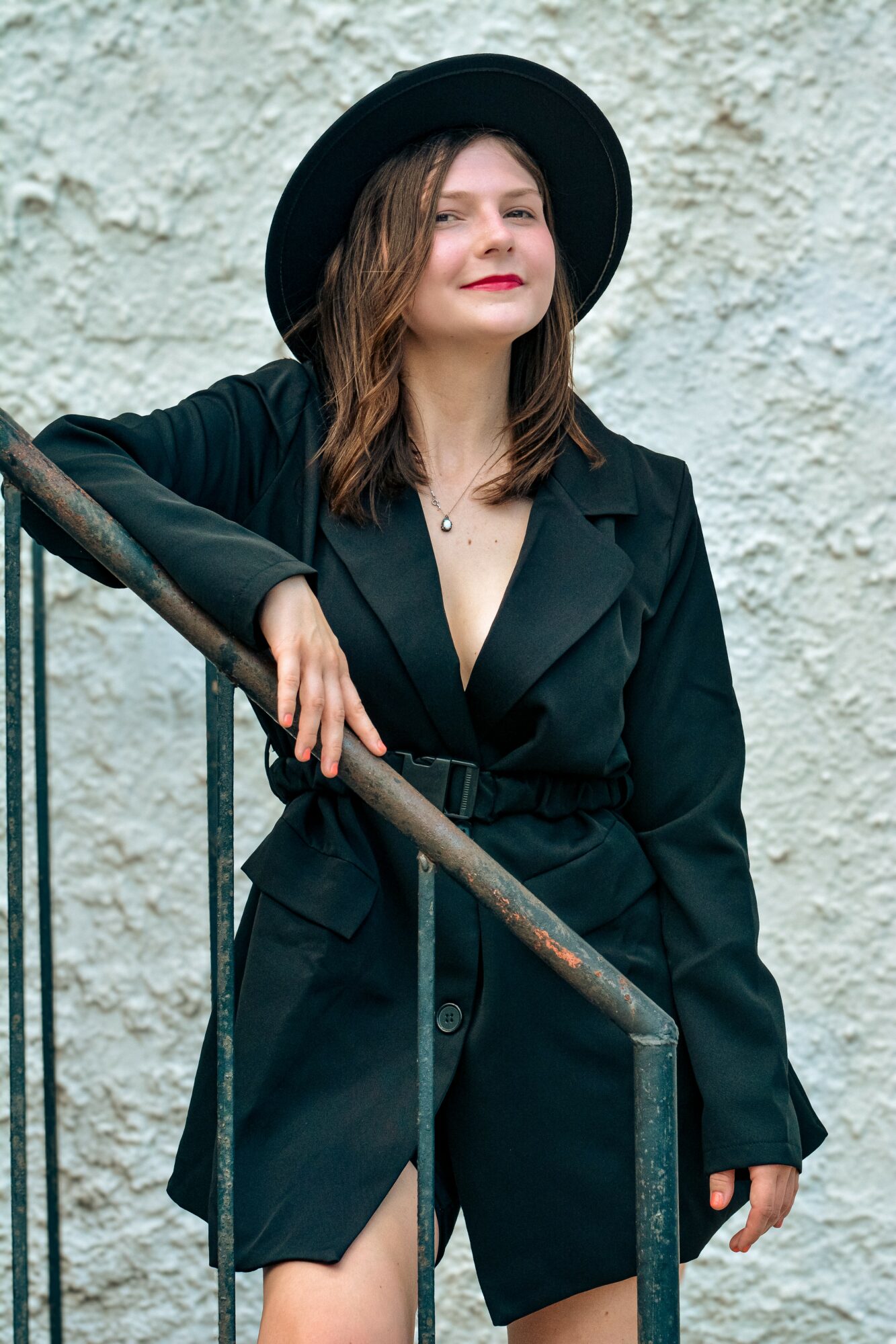 Woman with shoulder-length hair wearing a black hat and coat, standing outdoors against a textured wall.