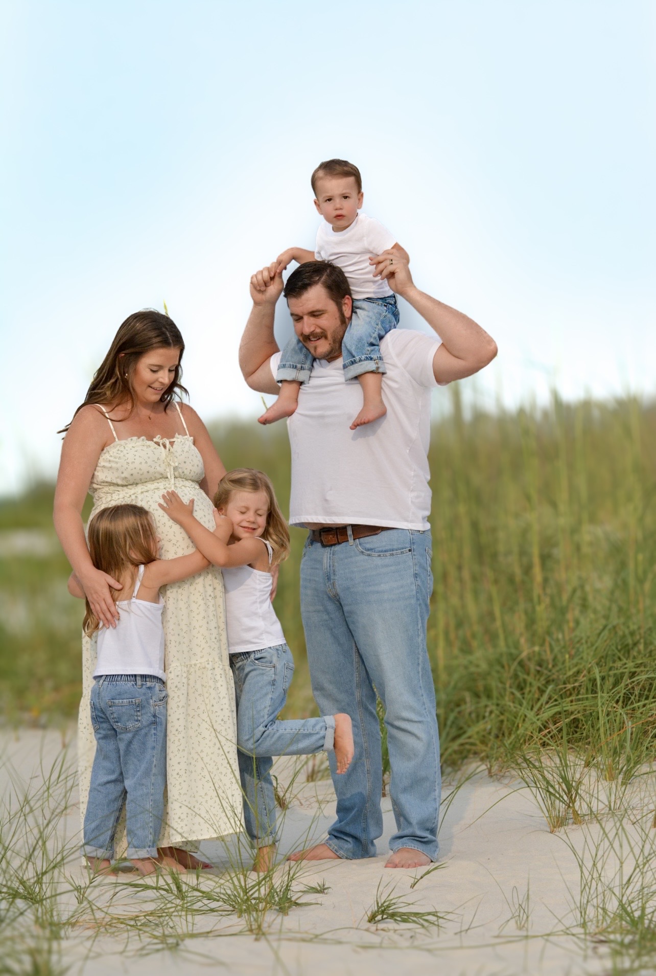 Family outdoors on a sandy area with grass, a man carrying a child on his shoulders, and two women with children, smiling.