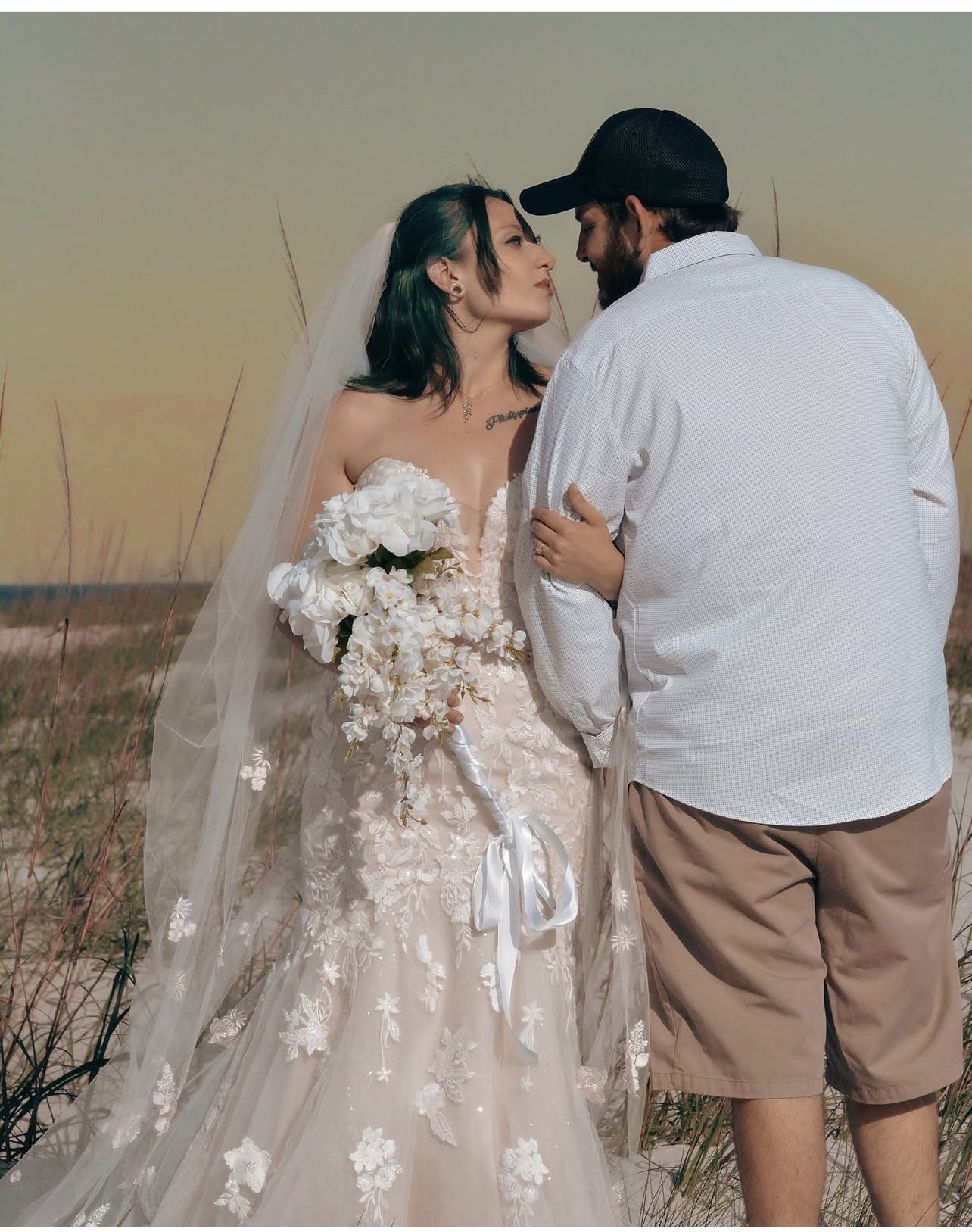 A bride and groom stand close together outdoors at sunset, gazing at each other, with a field in the background.