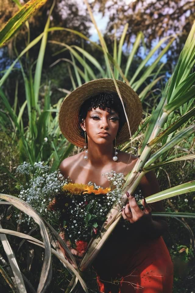 Woman with short curly hair wearing a wide-brimmed hat, surrounded by tall green plants, holding a flower bouquet.