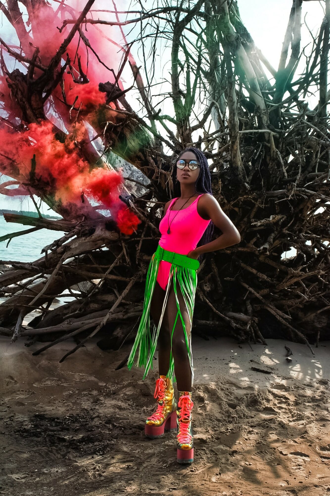 Woman in pink top and green skirt with long green fringe, standing on beach with tangled tree roots behind her.