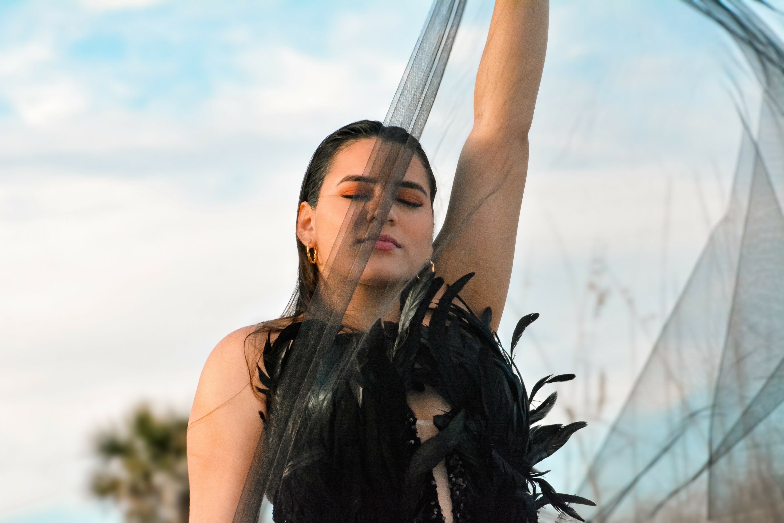 Woman with eyes closed, raising her arm, outdoors with sky and trees in background.