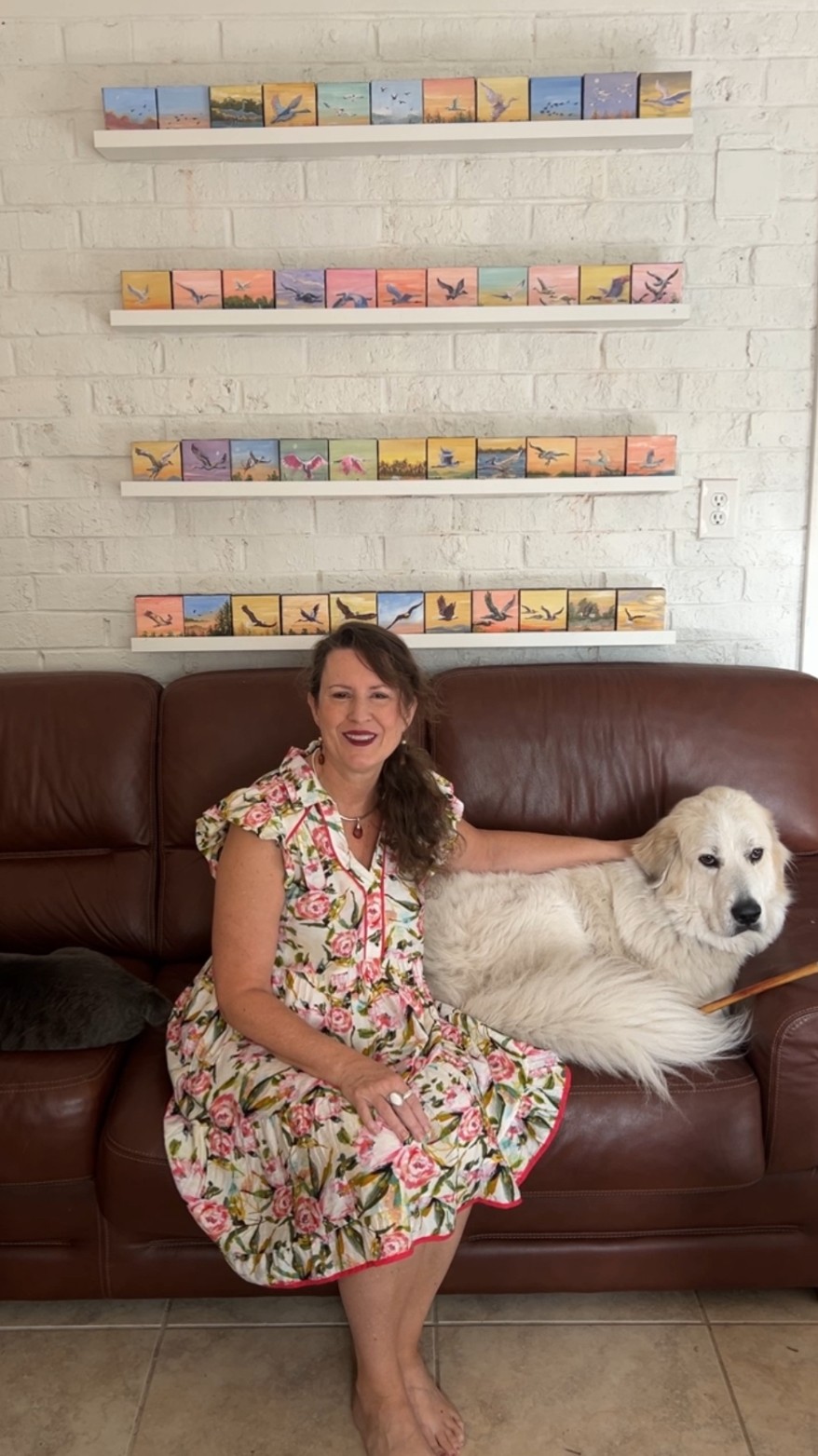 Woman in floral dress sitting on sofa with large white dog, brick wall with colorful artwork behind them.