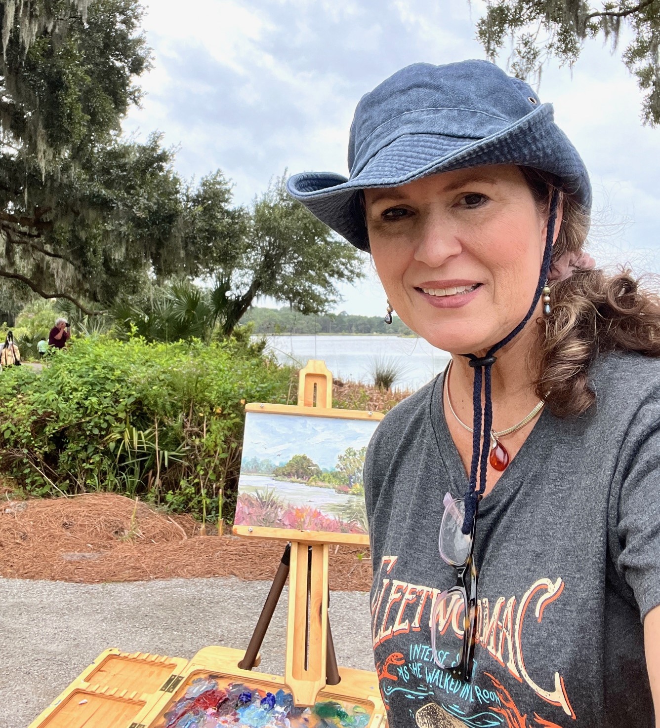 Woman wearing a blue hat and gray T-shirt outdoors near water and trees, smiling at the camera.