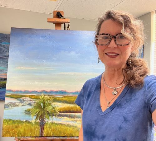 Woman with curly hair and glasses smiling in front of a landscape painting of a lake, palm tree, and sky.