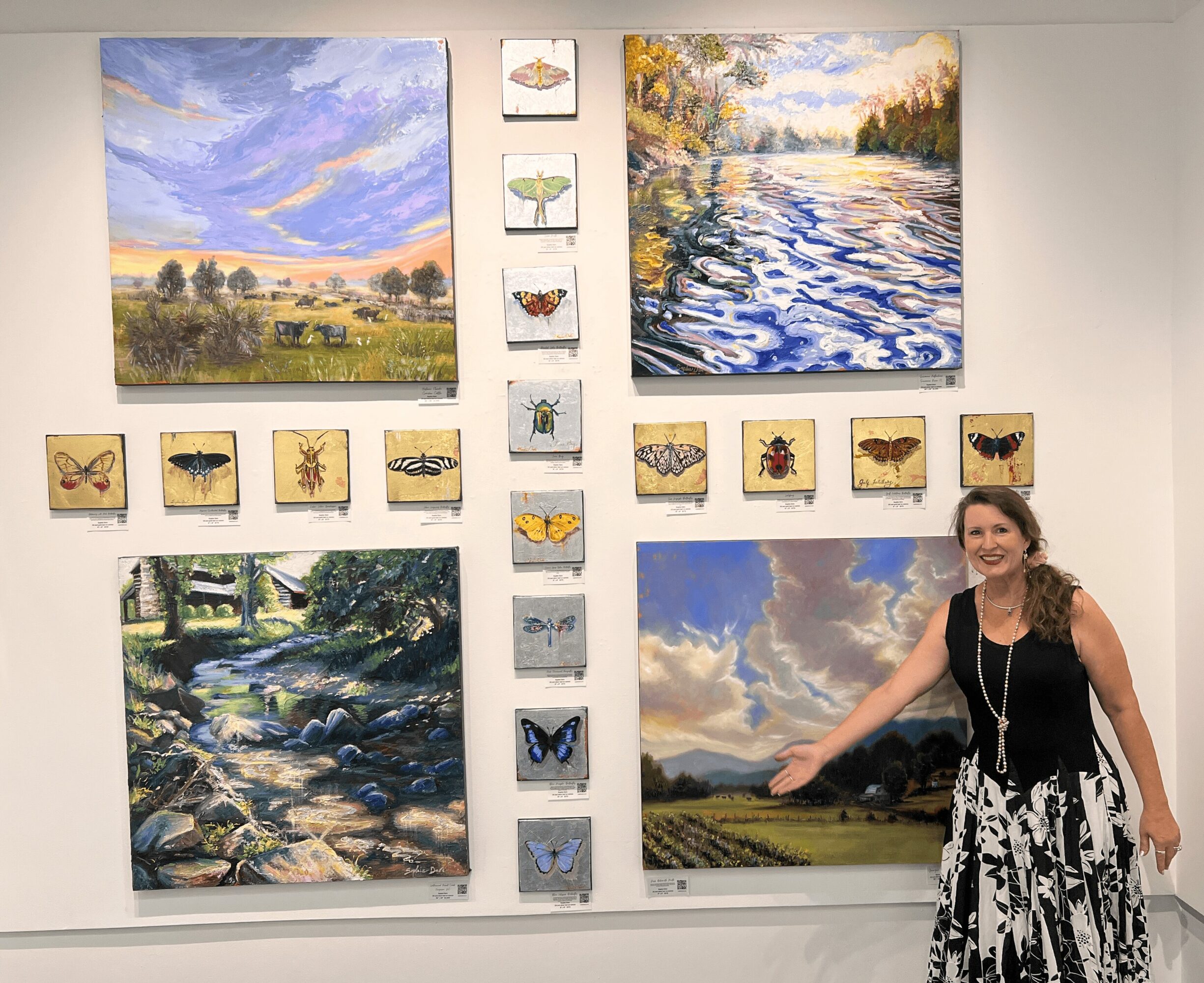 Woman standing in front of a wall with landscape and butterfly paintings, smiling and gesturing towards the artwork.