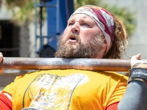 Man with a beard and headband lifting a barbell, wearing a yellow shirt, outdoors with blurred background.