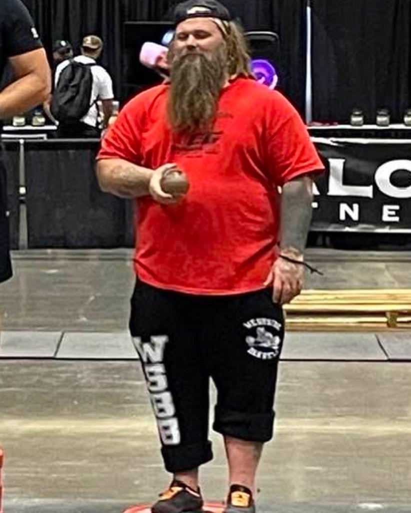Bearded man in red shirt and black shorts standing indoors, holding a small object, with people and equipment in background.