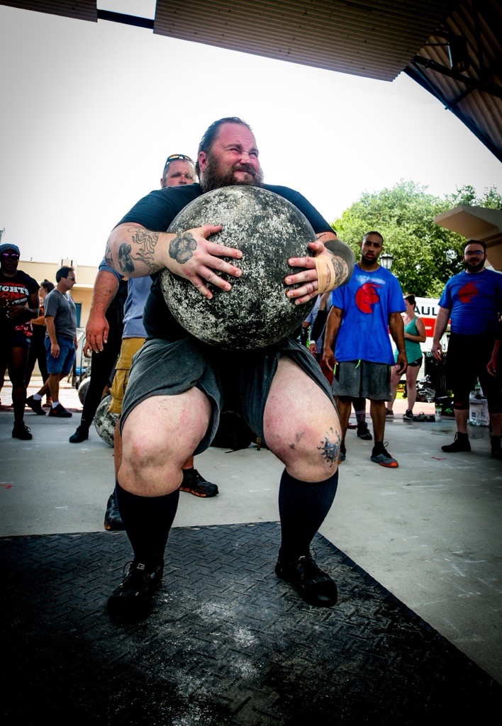Man lifting a large stone during a strongman event, surrounded by onlookers outdoors.