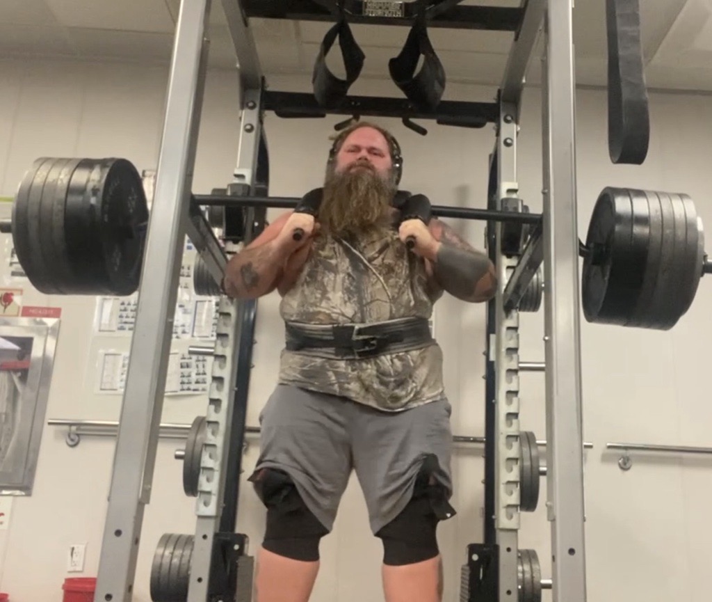 Man with a beard lifting a barbell in a gym, wearing a camouflage shirt and shorts, with gym equipment in background.