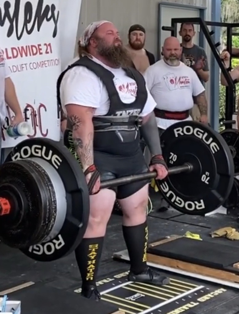 A man with a beard and tattoos lifts a heavy barbell in a gym, with three people watching behind him.