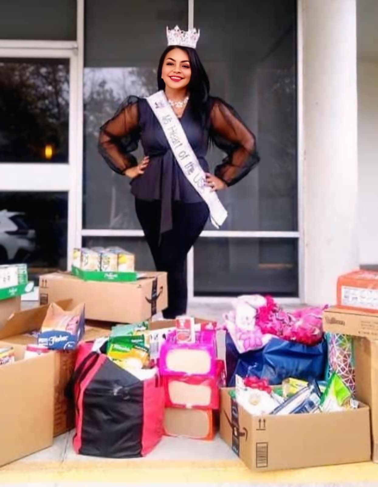 Woman wearing a crown and sash, standing behind boxes of gifts, smiling outdoors.