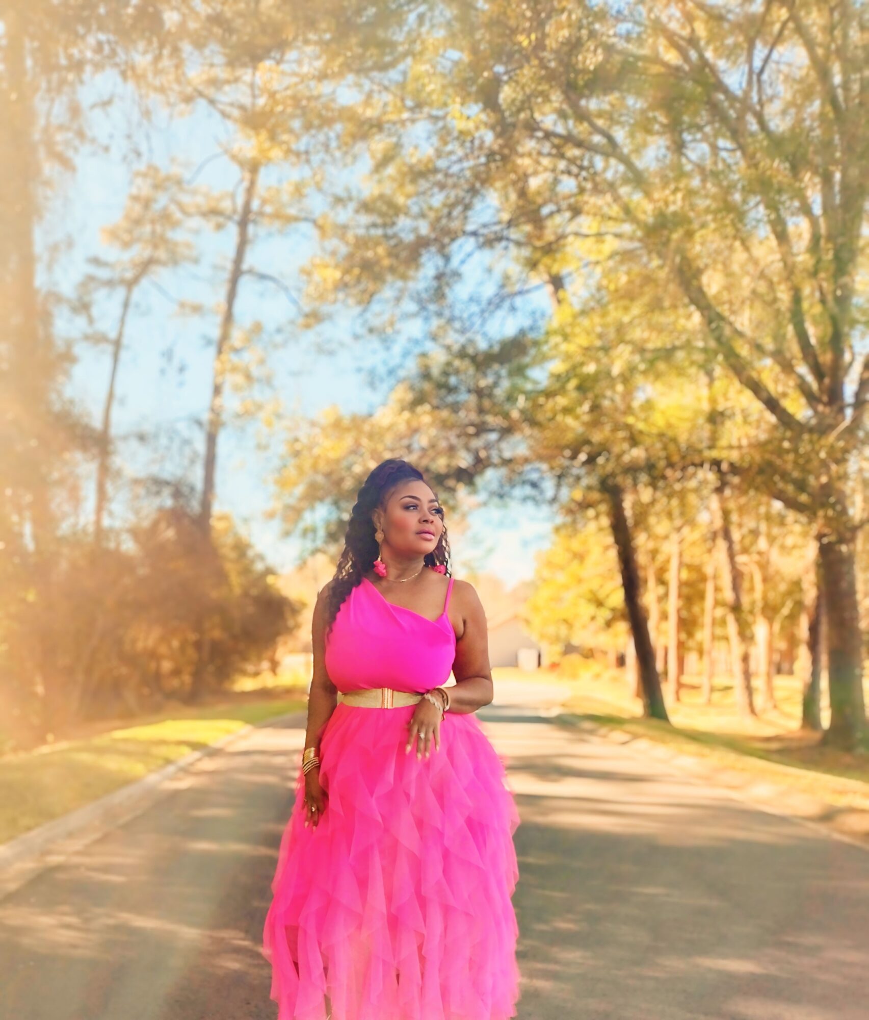 Woman in pink dress standing on a tree-lined path during autumn, sunlight filtering through leaves.