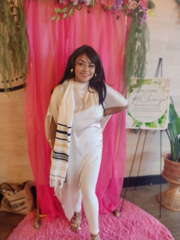Young woman with long dark hair smiling, standing in front of pink curtain with floral decorations, wearing white outfit.