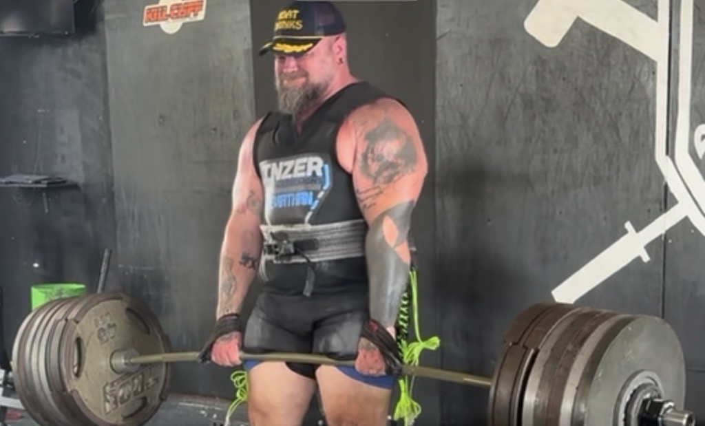 A muscular man with tattoos lifting a heavy barbell in a gym, wearing a tank top, cap, and glasses.