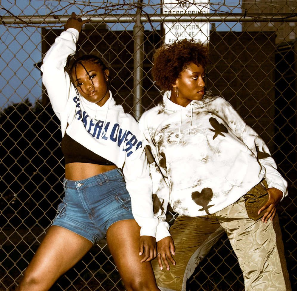 Two women stand in front of a chain-link fence, one with arm raised, both looking to the side, urban background.