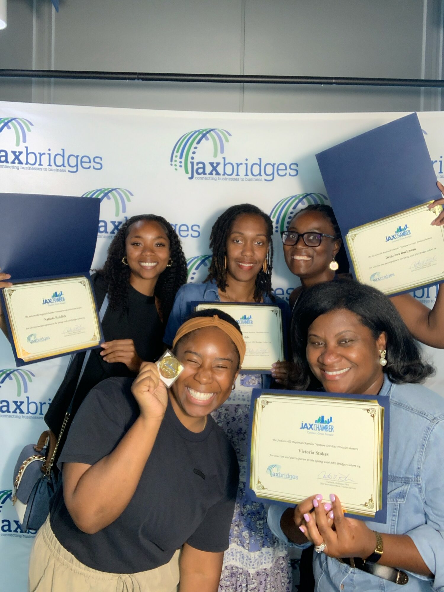 Five women holding certificates and a medal, smiling in front of a Jaxbridges backdrop.