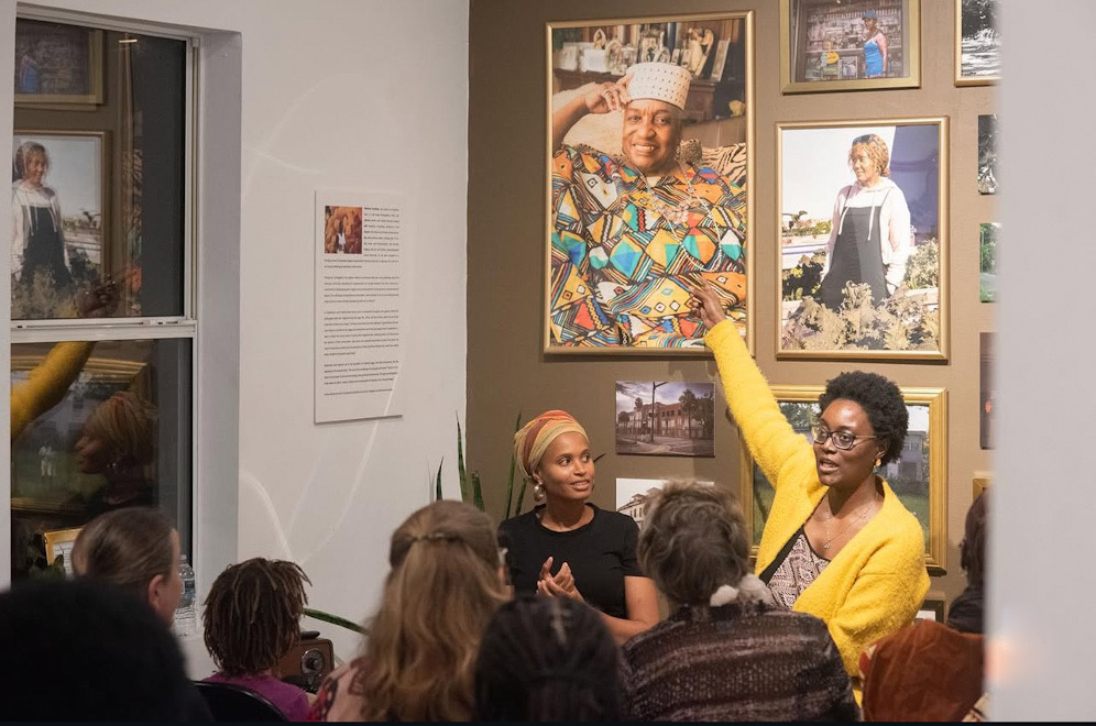 Two women speaking to an audience in an art gallery, one gesturing towards framed photographs on the wall.