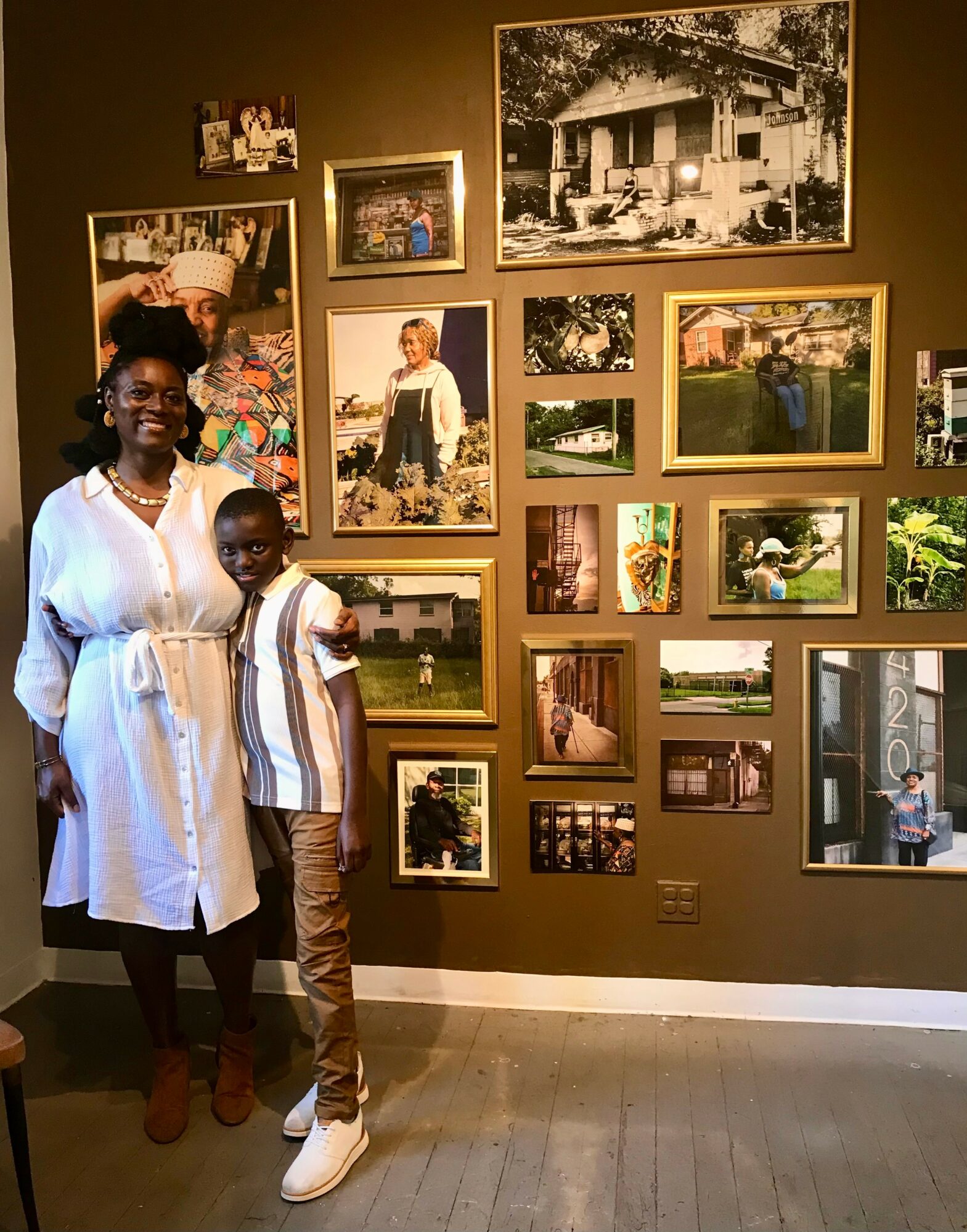 Two people, a woman and a child, stand in front of a wall with framed photographs, smiling at the camera.