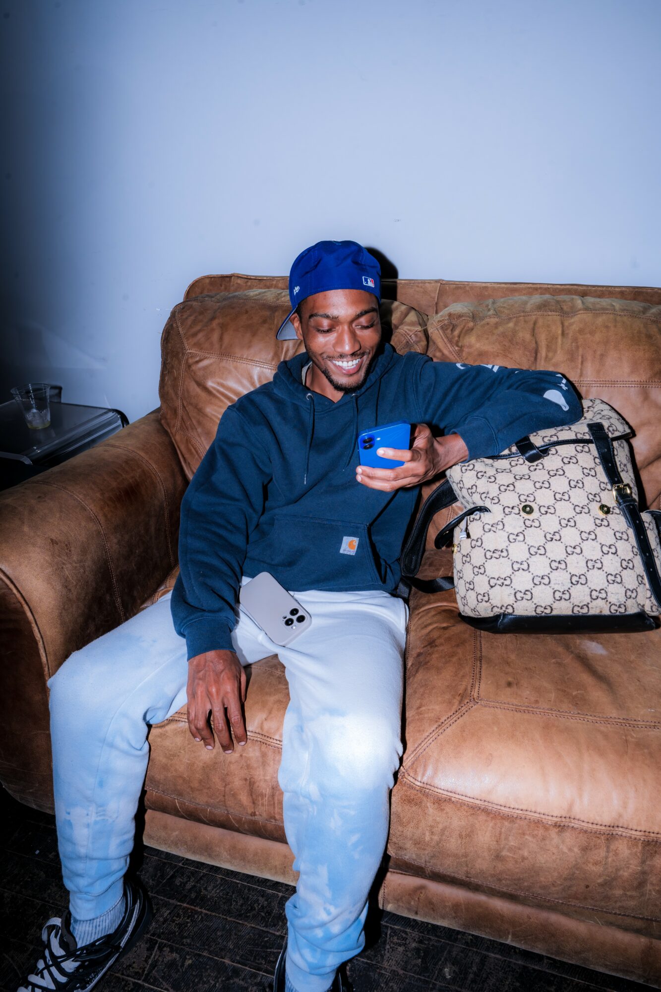 Smiling man in blue cap and jacket sitting on brown sofa, looking at phone, with a patterned bag beside him.