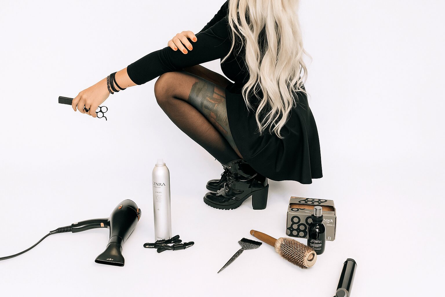 Person with long hair squatting, holding scissors, surrounded by hair styling tools and products, against a white background.